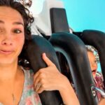 Woman on a roller coaster giving a thumbs-up, smiling at the camera with a blue background behind her.