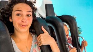 Woman on a roller coaster giving a thumbs-up, smiling at the camera with a blue background behind her.