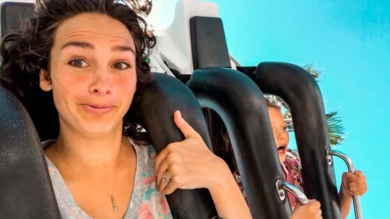 Woman on a roller coaster giving a thumbs-up, smiling at the camera with a blue background behind her.
