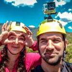 Two hikers wearing yellow helmets take a selfie outdoors, with a GoPro mounted on one helmet and mountain scenery behind them.