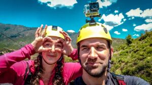 Two hikers wearing yellow helmets take a selfie outdoors, with a GoPro mounted on one helmet and mountain scenery behind them.