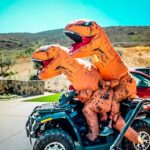 Two people in orange inflatable dinosaur costumes riding an ATV in a sunny residential driveway.