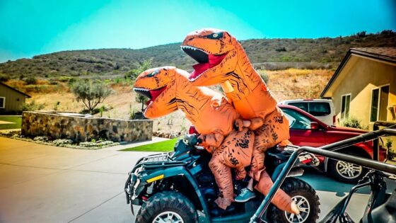 Two people in orange inflatable dinosaur costumes riding an ATV in a sunny residential driveway.