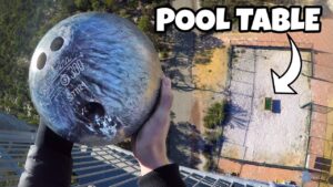 Person grasps a bowling ball from a high balcony, looking down on a courtyard with a small pool table and the words 'POOL TABLE' marked by an arrow.