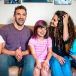 A family of five (father, mother, and three daughters) sitting on a brown couch at home, smiling together in a living room.