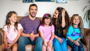 A family of five (father, mother, and three daughters) sitting on a brown couch at home, smiling together in a living room.