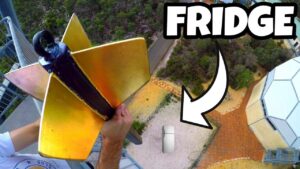 YouTube thumbnail showing a person on a high platform holding a yellow winged contraption, with a fridge on the ground below and the word FRIDGE in bold text.