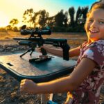 Girl in a floral dress smiles while using a scoped rifle on a shooting rest in a dry field at sunset