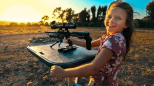 Girl in a floral dress smiles while using a scoped rifle on a shooting rest in a dry field at sunset
