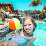 Smiling girl in a swimming pool by the edge with a colorful beach ball nearby on a sunny day at a tropical resort.