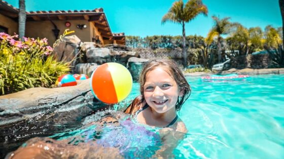 Smiling girl in a swimming pool by the edge with a colorful beach ball nearby on a sunny day at a tropical resort.