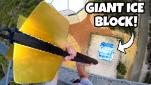 Drone‑shot thumbnail of a person on a tall tower, gripping a large gold tool toward a blue ice block on the ground far below; bold caption reads 'GIANT ICE BLOCK!' indicating a stunt video.
