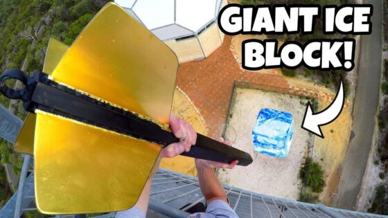 Drone‑shot thumbnail of a person on a tall tower, gripping a large gold tool toward a blue ice block on the ground far below; bold caption reads 'GIANT ICE BLOCK!' indicating a stunt video.