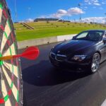 Black BMW convertible on a drag strip with a large dartboard in the foreground, a red dart near the bullseye as the car approaches.