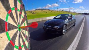 Black BMW convertible on a drag strip with a large dartboard in the foreground, a red dart near the bullseye as the car approaches.