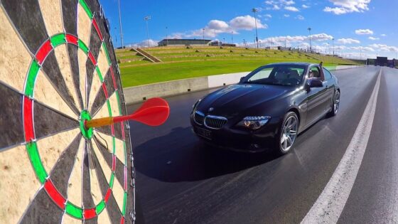 Black BMW convertible on a drag strip with a large dartboard in the foreground, a red dart near the bullseye as the car approaches.
