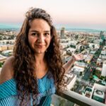Smiling woman with long curly hair on a rooftop balcony, overlooking a city at sunset with arms open wide.