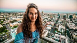 Smiling woman with long curly hair on a rooftop balcony, overlooking a city at sunset with arms open wide.
