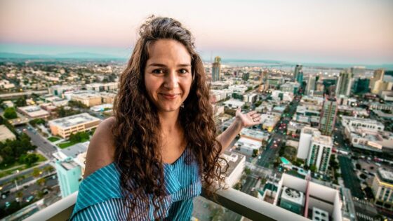 Smiling woman with long curly hair on a rooftop balcony, overlooking a city at sunset with arms open wide.