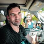 Man in a black shirt sits in the driver's seat of a car, looking at the camera while two people stand outside smiling at the window.