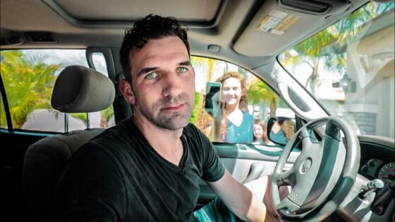 Man in a black shirt sits in the driver's seat of a car, looking at the camera while two people stand outside smiling at the window.