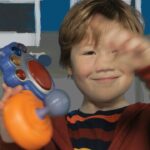 Smiling young boy plays with a colorful toy controller indoors, waving hello.