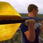 Man wearing sunglasses on a railing holds a large yellow arrow-shaped object toward the camera, overlooking a forested landscape.