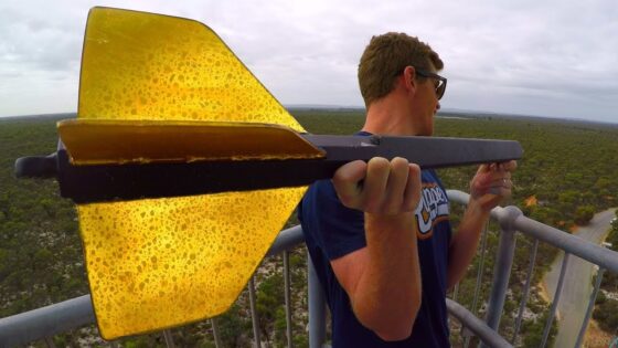 Man wearing sunglasses on a railing holds a large yellow arrow-shaped object toward the camera, overlooking a forested landscape.