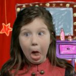 Girl on a playful stage backdrop making a goofy, surprised face with tongue out. Behind her is a red curtain and a cartoon dressing table with lights and stars.