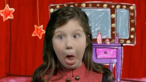 Girl on a playful stage backdrop making a goofy, surprised face with tongue out. Behind her is a red curtain and a cartoon dressing table with lights and stars.