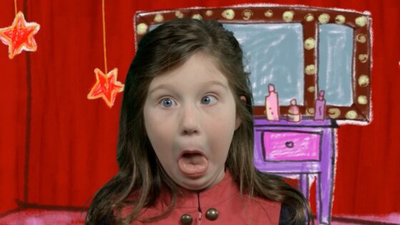 Girl on a playful stage backdrop making a goofy, surprised face with tongue out. Behind her is a red curtain and a cartoon dressing table with lights and stars.