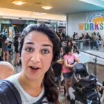 Woman wearing a baby carrier takes a selfie in a crowded mall near Build-A-Bear Workshop sign.