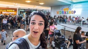 Woman wearing a baby carrier takes a selfie in a crowded mall near Build-A-Bear Workshop sign.