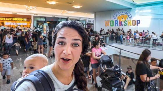 Woman wearing a baby carrier takes a selfie in a crowded mall near Build-A-Bear Workshop sign.