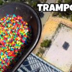 Aerial view of a large tub filled with rainbow jelly beans beside a balcony railing, overlooking a fenced outdoor area with a square trampoline below.