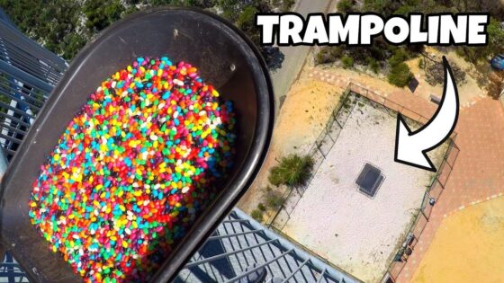 Aerial view of a large tub filled with rainbow jelly beans beside a balcony railing, overlooking a fenced outdoor area with a square trampoline below.