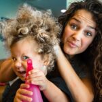Woman with long curly hair playfully styles a child’s wild curls as the child holds a pink spray bottle at the shop.
