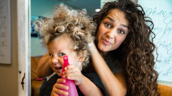 Woman with long curly hair playfully styles a child’s wild curls as the child holds a pink spray bottle at the shop.