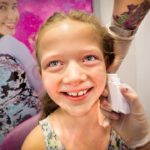 Smiling young girl in a store as a gloved professional uses a white device on her cheek, with colorful product displays in the background.