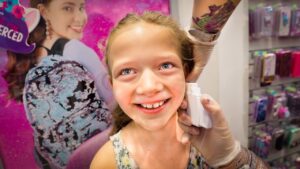 Smiling young girl in a store as a gloved professional uses a white device on her cheek, with colorful product displays in the background.