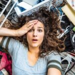 Woman lying on her back amid a chaotic pile of belongings, holding her forehead in surprise or worry, eyes wide.