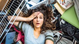 Woman lying on her back amid a chaotic pile of belongings, holding her forehead in surprise or worry, eyes wide.