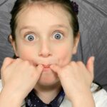 Young girl with wide blue eyes making a silly face, pulling the corners of her mouth with her fists.