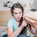 Young woman indoors holds a long wooden plank, leaning on it while looking at the camera in a living room setting with photos on the wall behind her.