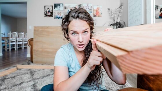Young woman indoors holds a long wooden plank, leaning on it while looking at the camera in a living room setting with photos on the wall behind her.