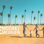 Family of six walking hand-in-hand across a sandy hill under tall palm trees, with large 'The Moore Merrier' text on the left (banner photo).
