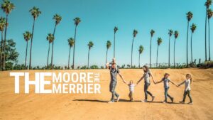 Family of six walking hand-in-hand across a sandy hill under tall palm trees, with large 'The Moore Merrier' text on the left (banner photo).