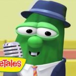 Green VeggieTales character wearing a blue fedora and suit, singing into a silver microphone with a colorful stage backdrop and VeggieTales logo in the corner.