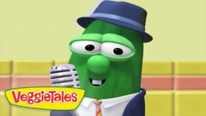Green VeggieTales character wearing a blue fedora and suit, singing into a silver microphone with a colorful stage backdrop and VeggieTales logo in the corner.