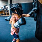 Young girl wearing boxing headgear and gloves punches a hanging bag while a coach holds it for guidance in a gymenario setting.